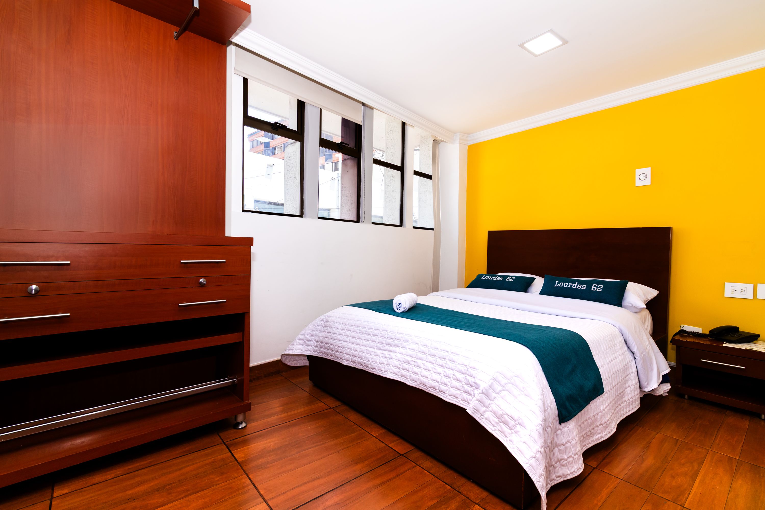 A hotel room with a wooden dresser, a bed with a white quilt, and a yellow accent wall.