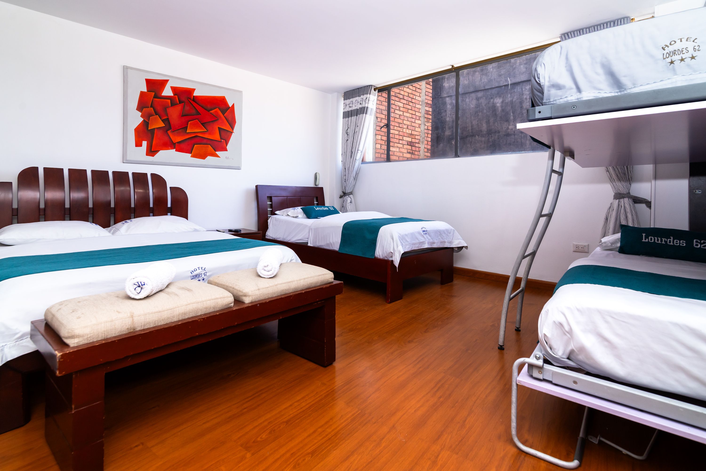 A hotel room with two beds, a bench, a window with a view of a brick wall, and a red abstract painting on the wall.