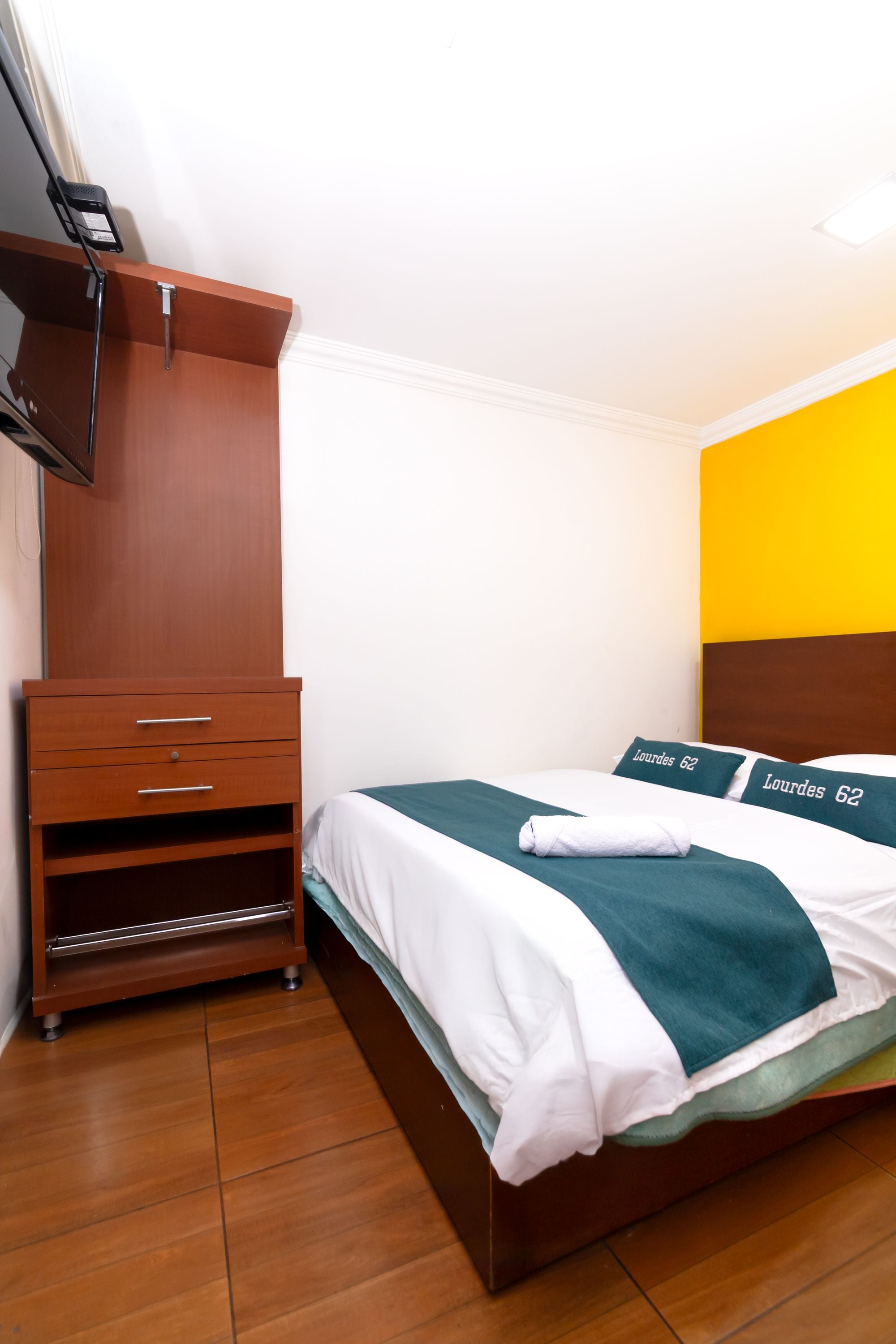 Habitación de hotel con cómoda de madera, cama con ropa de cama verde y blanca, y una pared de acento amarilla.