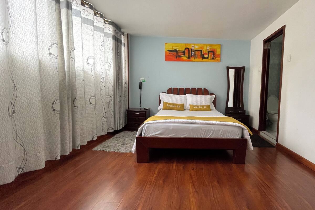 Un dormitorio con una cama de madera, ropa de cama blanca, un cojín amarillo decorativo, un cuadro en la pared y piso de madera.