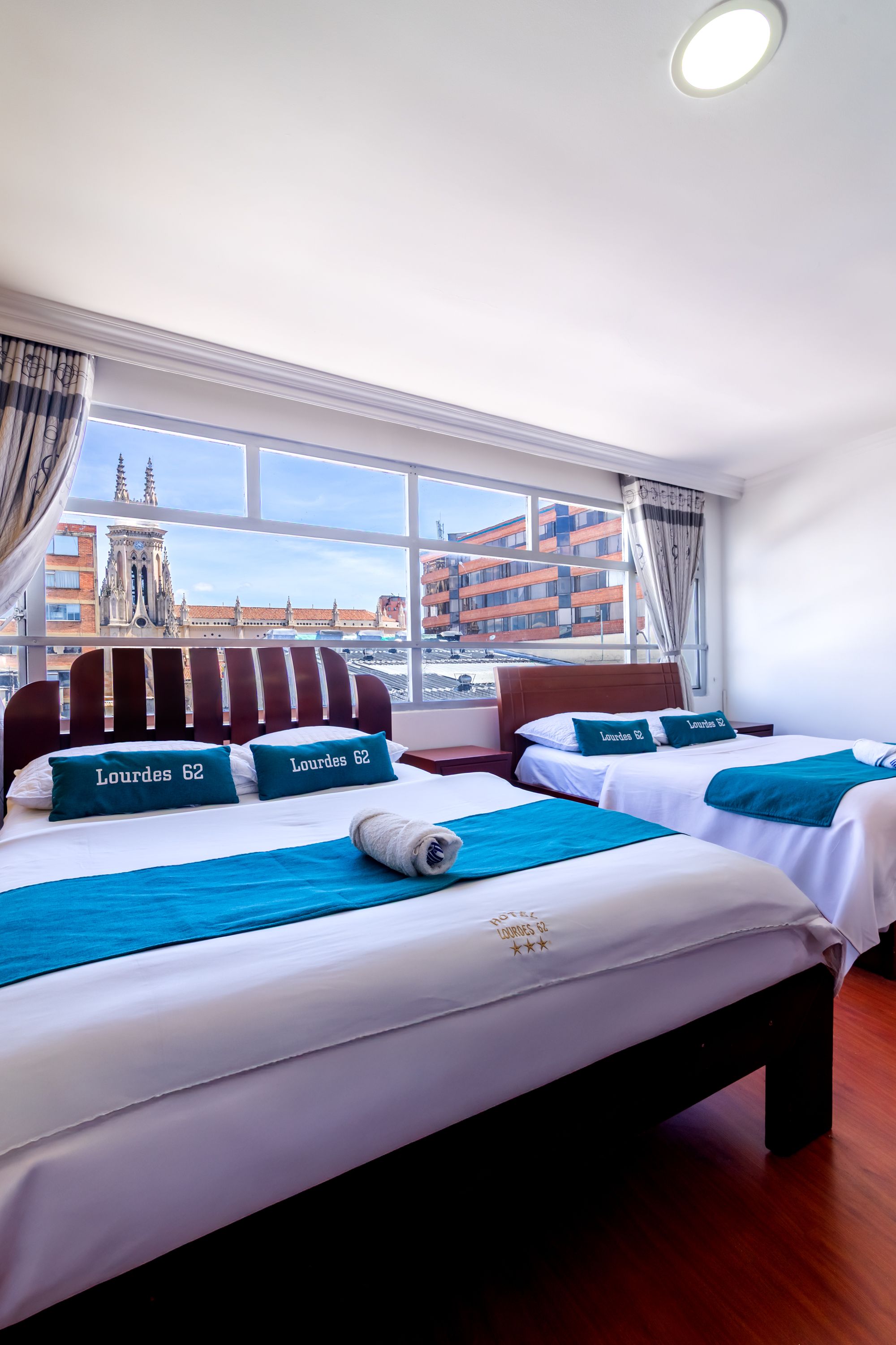 Habitación de hotel con dos camas, cada una con cubrecama y almohadas con la etiqueta 'Lourdes 62'. La habitación tiene ventanas amplias que ofrecen vista a un paisaje urbano con una iglesia y edificios modernos.