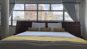 Ein Hotelzimmer mit einem großen Bett, drei Kissen mit dem Aufdruck "Lourdes 62", einem hölzernen Kopfteil und Blick auf Gebäude durch große Fenster.