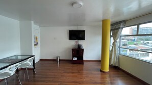 A room with a wooden floor, a glass table with chairs, a TV mounted on the wall, a small cabinet, and a window with a view of buildings. - Hotel Lourdes 62 (Bogotá)