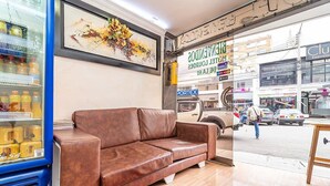 A modern interior with a brown leather sofa, a blue refrigerator stocked with beverages, and a large window overlooking a busy street. - Hotel Lourdes 62 (Bogotá)