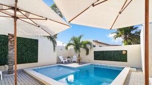 Una piscina rectangular con solárium empinado, rodeada de sillas y sombrillas blancas, palmeras y un muro verde.