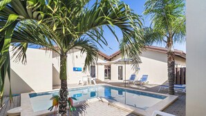 A swimming pool with lounge chairs, palm trees, and a building with a tiled roof. - Aruba 1BR Apartment w Free Parking near Palm Beach (Noord)
