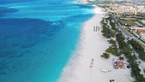 A coastal beach area with a white sandy shore, clear turquoise waters, and a row of buildings along the beachfront. - Aruba 1BR Apartment w Free Parking near Palm Beach (Noord)