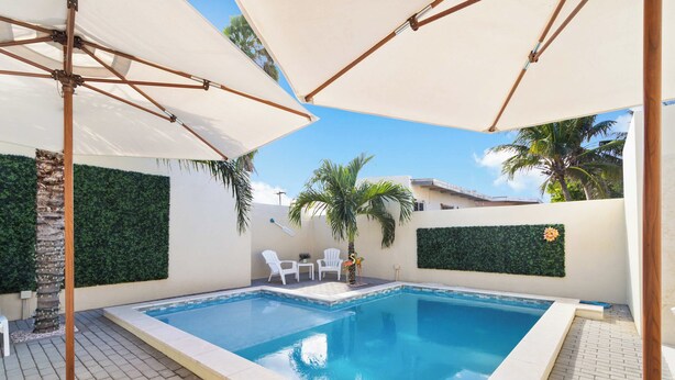 A rectangular swimming pool with a tiled edge, surrounded by a patio area with white chairs and umbrellas. - Little Seas #3 Aruba 1BR Apartment w Free Parking near Palm Beach (Noord)