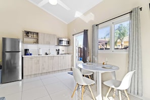 A modern kitchen with a refrigerator, microwave, and coffee maker, a dining area with a table and chairs, and a window with curtains. - Little Seas #3 Aruba 1BR Apartment w Free Parking near Palm Beach (Noord)