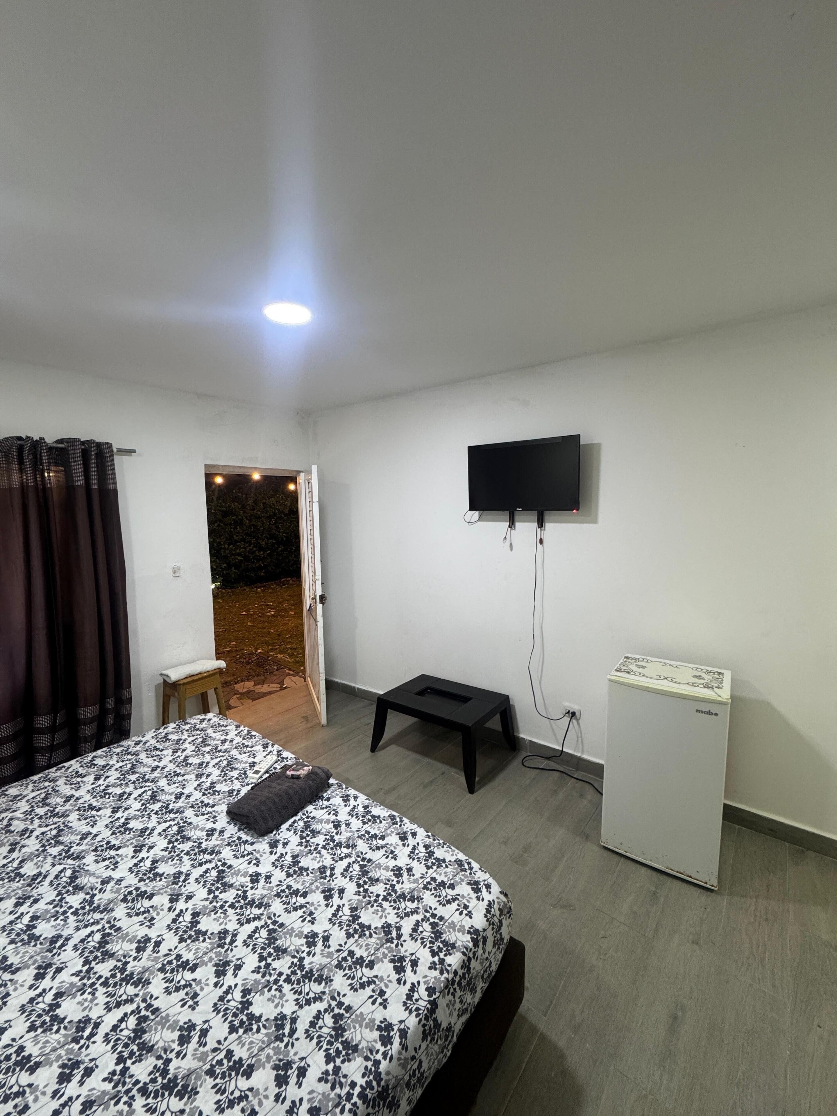 A bedroom with a bed, a small table, a television mounted on the wall, and a refrigerator.