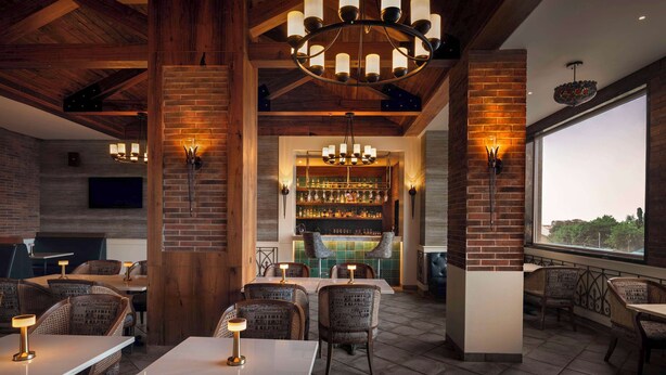 Un restaurante con paredes de ladrillo, vigas de madera y una zona de bar con estanterías llenas de botellas. Mesas y sillas dispuestas para comer.
