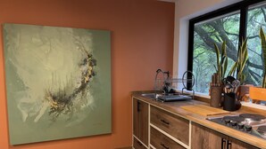 Ett modernt kök med en stor, abstrakt målning, träskåp, ett fönster med utsikt över grönområden och ett torkställ för disk.