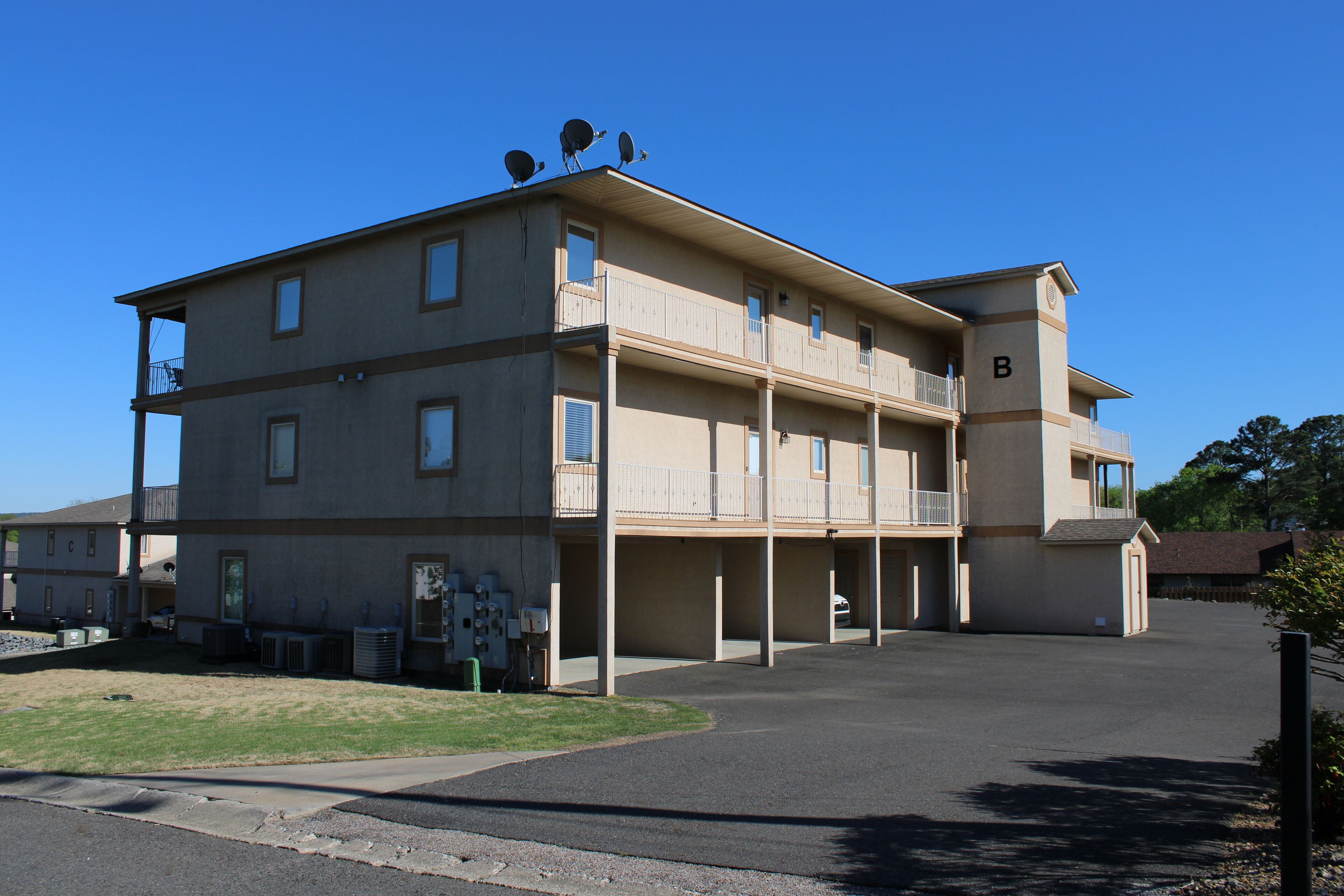 A multi-story building with a flat roof, balconies, and a parking area in front.