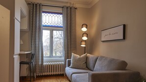 A modern living room with a grey sofa, a small table, and a chair. There is a window with patterned curtains, a radiator, and a wall-mounted light fixture.