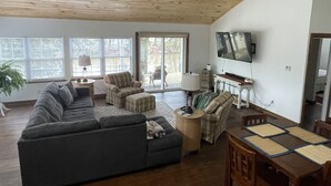 Un ampio soggiorno con un grande divano angolare grigio, un tavolo da pranzo in legno con quattro sedie, una ventola a soffitto e un televisore montato a parete.