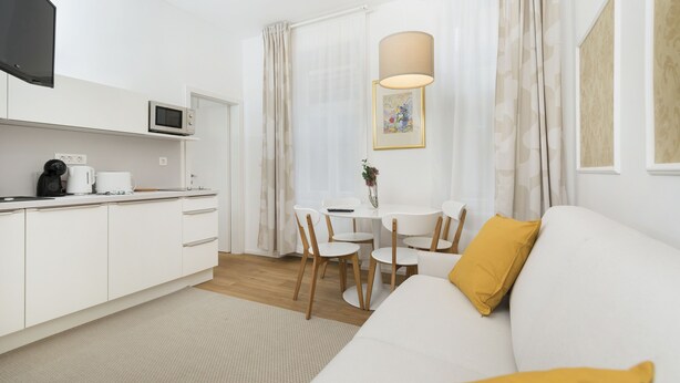 Une pièce moderne et bien éclairée, comprenant un coin cuisine, une table à manger avec des chaises et un canapé agrémenté d’un coussin jaune.