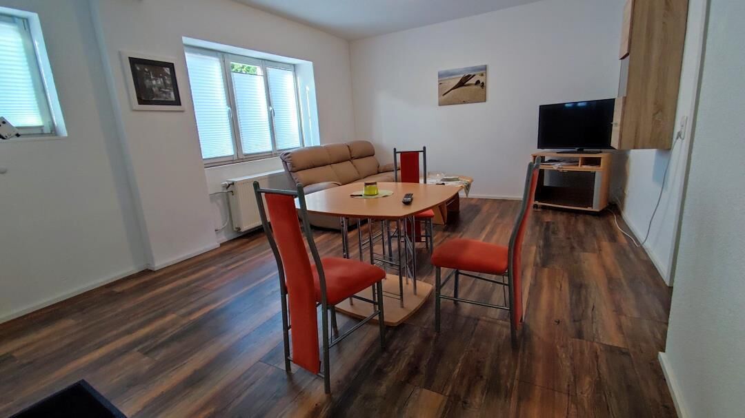 Ein Zimmer mit einem runden Tisch, vier Stühlen, einem Sofa, einem Fernseher auf einem Schrank und einem Fenster mit Jalousien.
