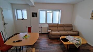 Ein Wohnzimmer mit braunem Sofa, einem kleinen Tisch mit Fernbedienung und einem Fenster mit Jalousien.