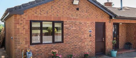 A small brick house with a tiled roof, a front door, and a window. There are potted plants and a hose on the ground.