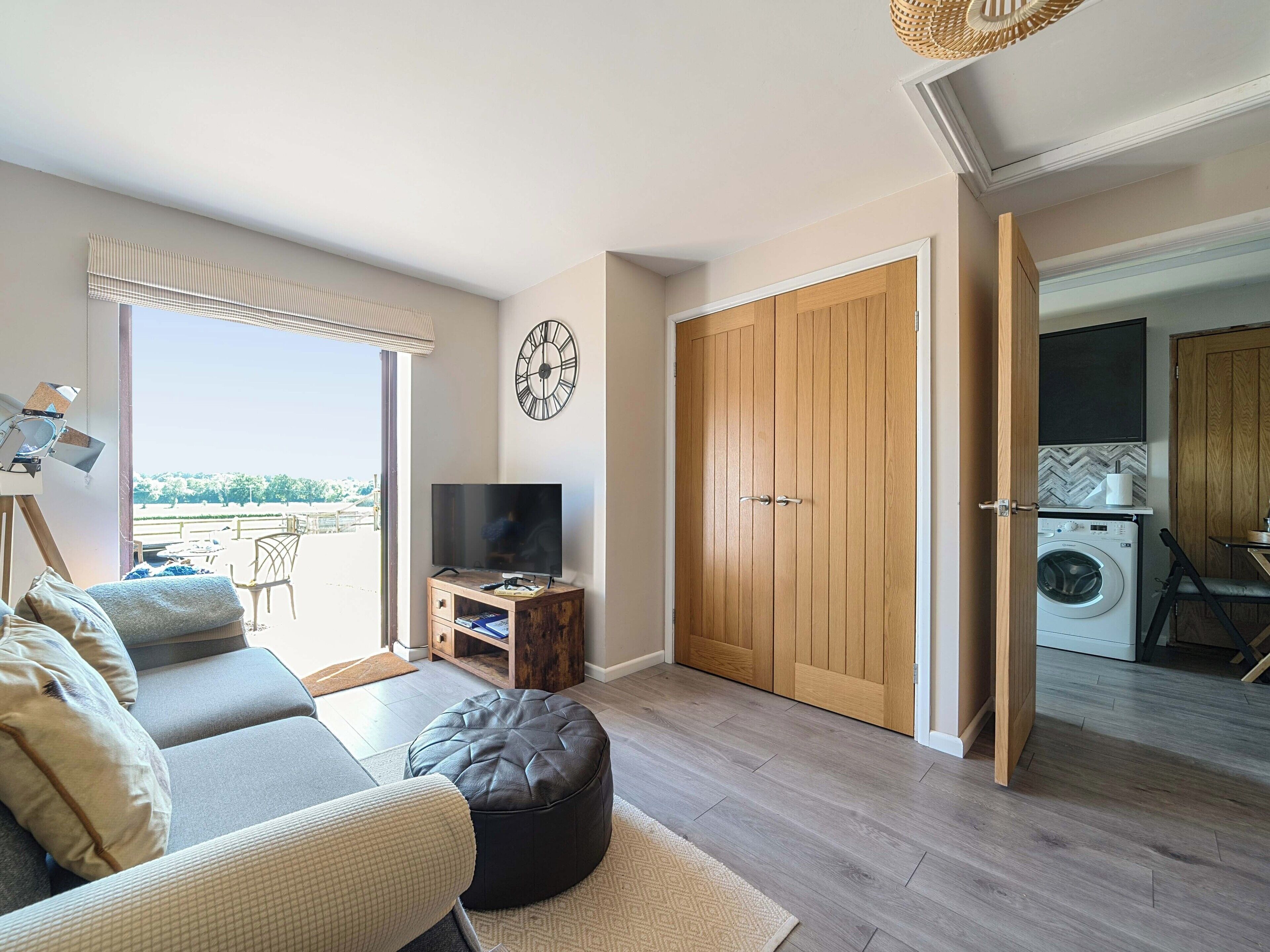 A modern living room with a sofa, ottoman, and a television on a wooden cabinet. A large wooden door leads to a laundry area.