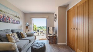 Ein Wohnzimmer mit grauem Sofa, Ottoman und einem Gemälde von Blumen. Ein Holzschrank und eine Tür zum Außenbereich sind ebenfalls vorhanden.