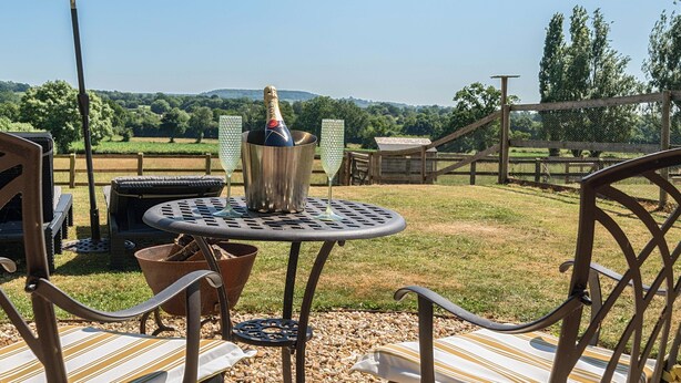 Eine Terrasse mit Gartenmöbeln, einem Tisch mit einer Champagnerflasche und Blick auf eine ländliche Landschaft.