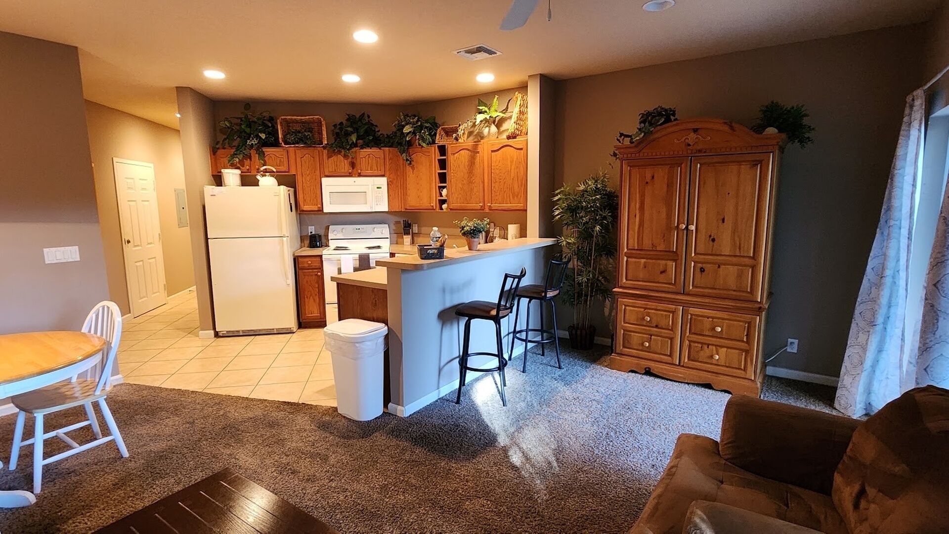 A kitchen with wooden cabinets, a refrigerator, a microwave, and a sink. There is a dining table with chairs and a bar area with stools.