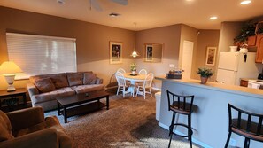 A living room with a sofa, coffee table, and dining table with chairs. There is a kitchen area with a refrigerator and cabinets.