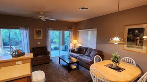A living room with a sofa, coffee table, dining table, and chairs. There is a ceiling fan, a window with curtains, and a lamp.