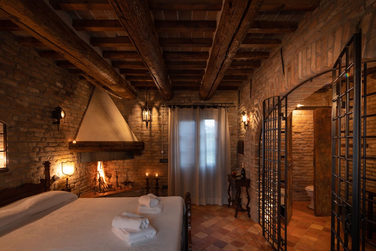 Una habitación rústica con vigas de madera a la vista, chimenea, cama con toallas ordenadas y vistas a través de una ventana con cortinas.