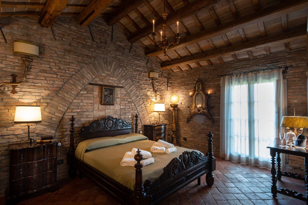 Una habitación tradicional con pared de piedra, techo de madera, cama con cabecera, mesitas de noche con lámparas y una ventana con cortinas.