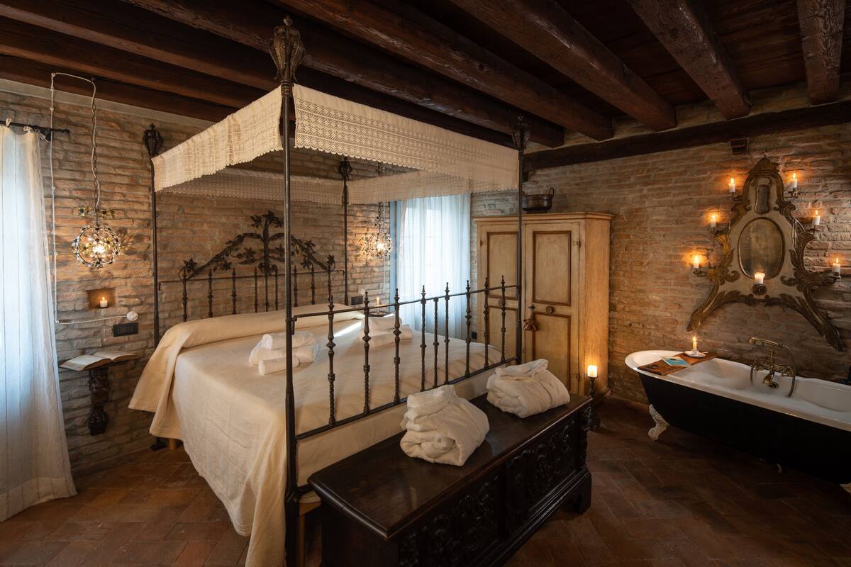 Un dormitorio tradicional con cama con dosel, un banco de madera con toallas, una bañera de patas de león y una pared de piedra.