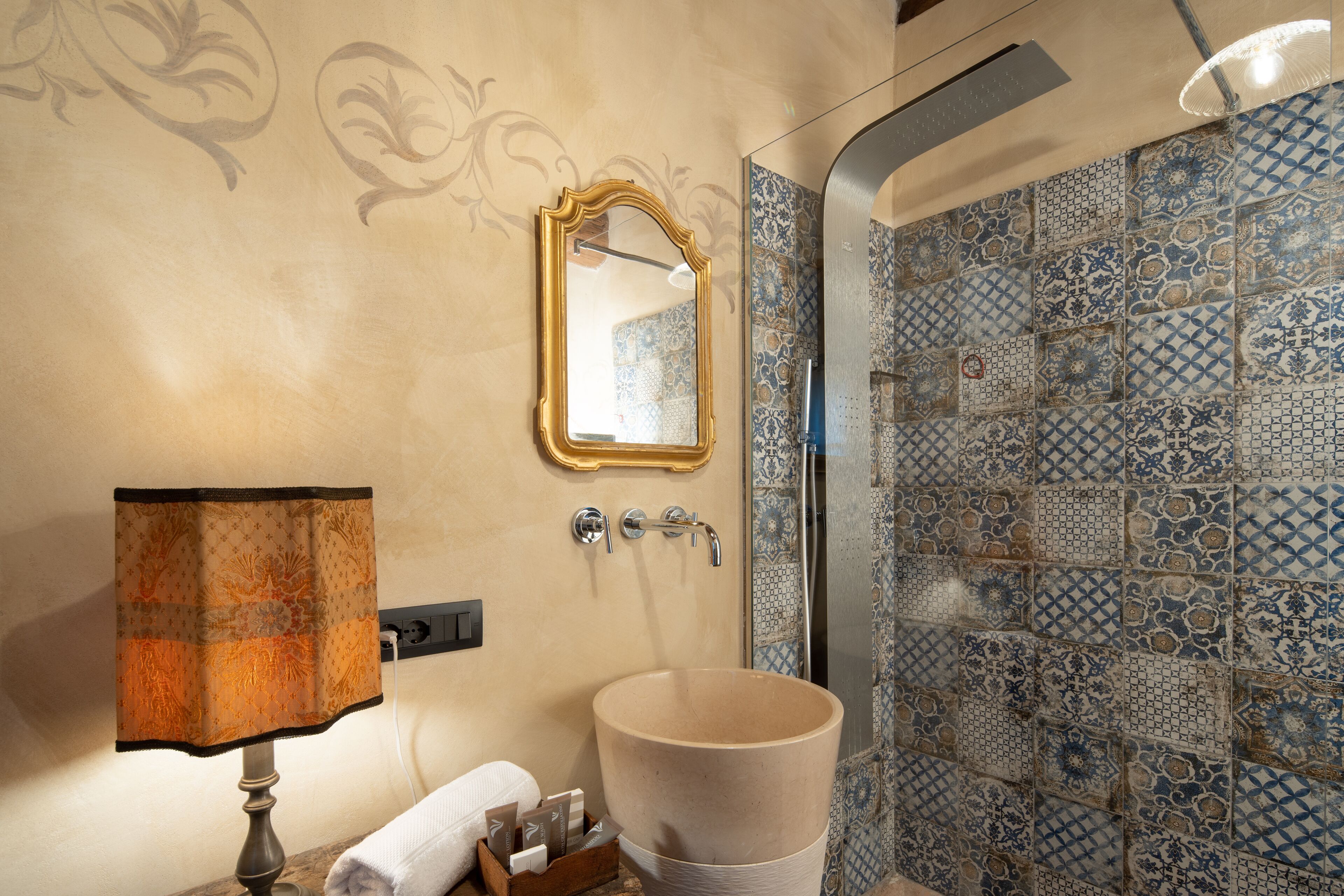 A bathroom with a beige sink, a mirror with a gold frame, and a patterned shower wall.