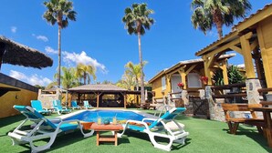 Un espace piscine avec des chaises longues, un parasol en chaume et un bâtiment à la façade jaune.