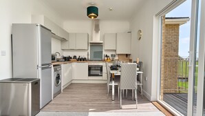 A modern kitchen with stainless steel appliances, a dining area with a table and chairs, and a view of a brick wall and greenery outside. - The Penthouse In Bonchurch  (Ventnor)