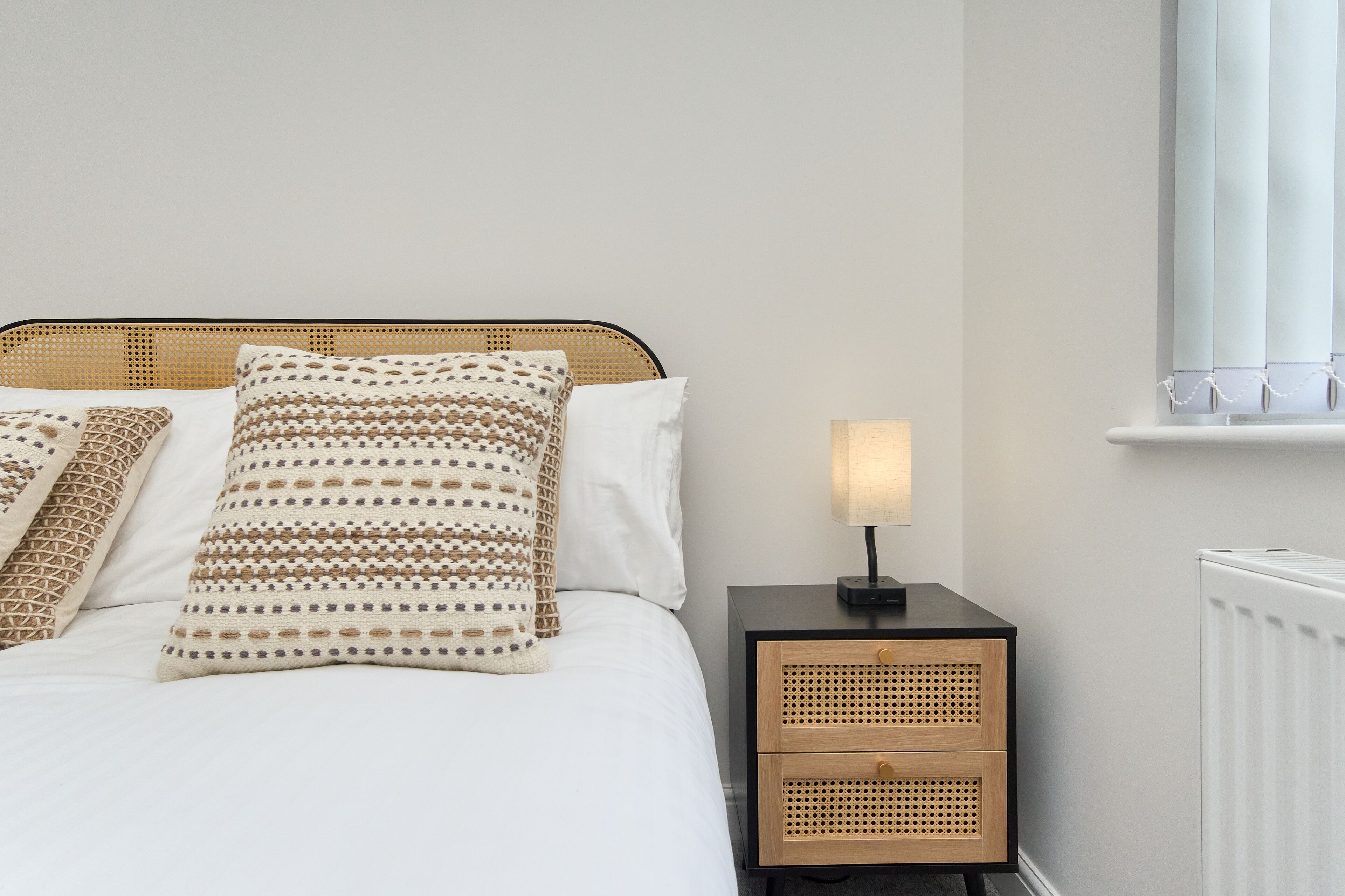 A neatly made bed with patterned pillows, a bedside table with a lamp, and a radiator.