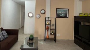 A living room with a brown sofa, a glass coffee table, a wall-mounted shelf with decorative items, a ceiling fan, and a framed picture on the wall.