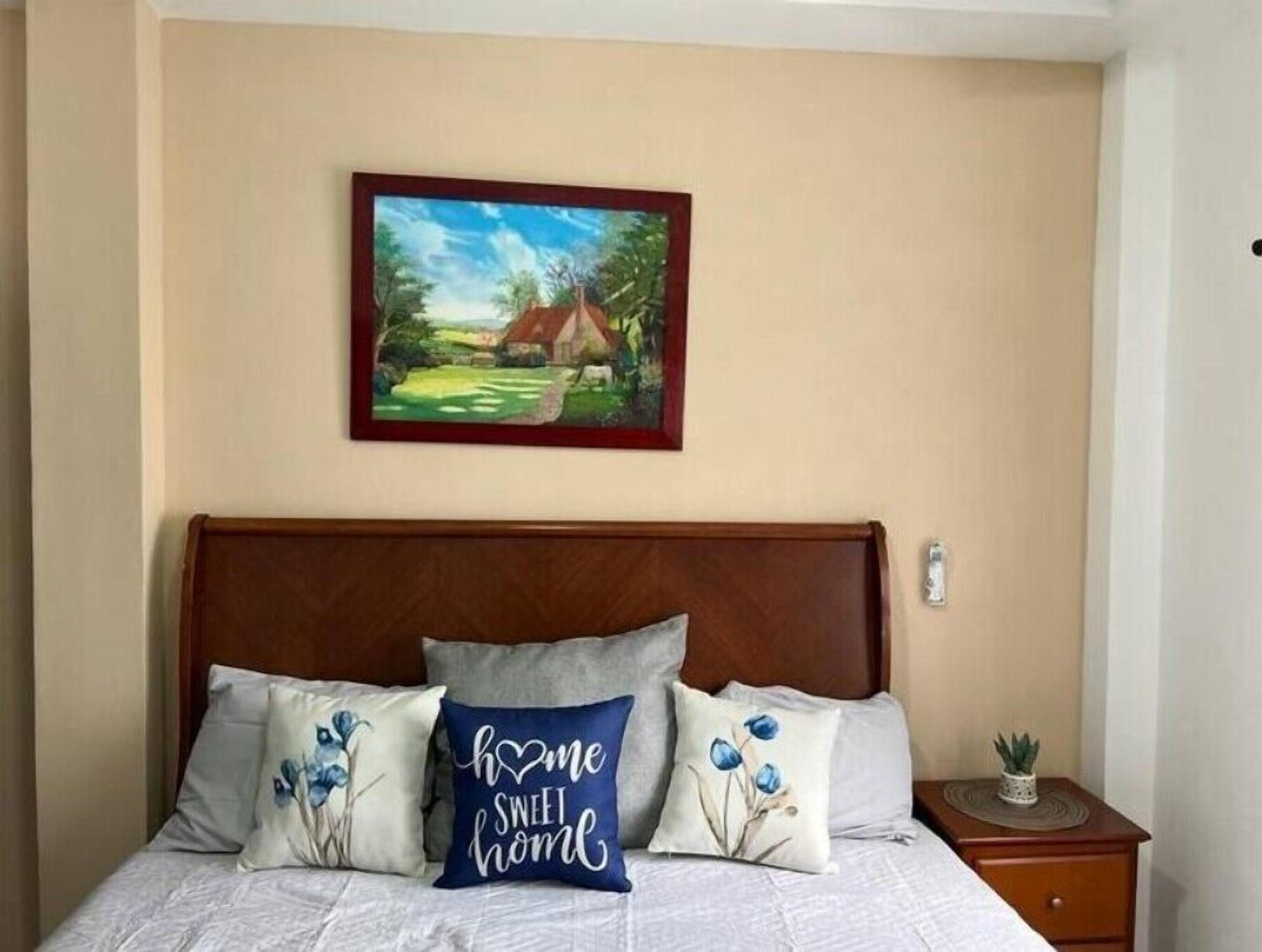 Une chambre à coucher avec un lit, des oreillers, une table de chevet, une petite plante en pot et un tableau encadré accroché au mur.