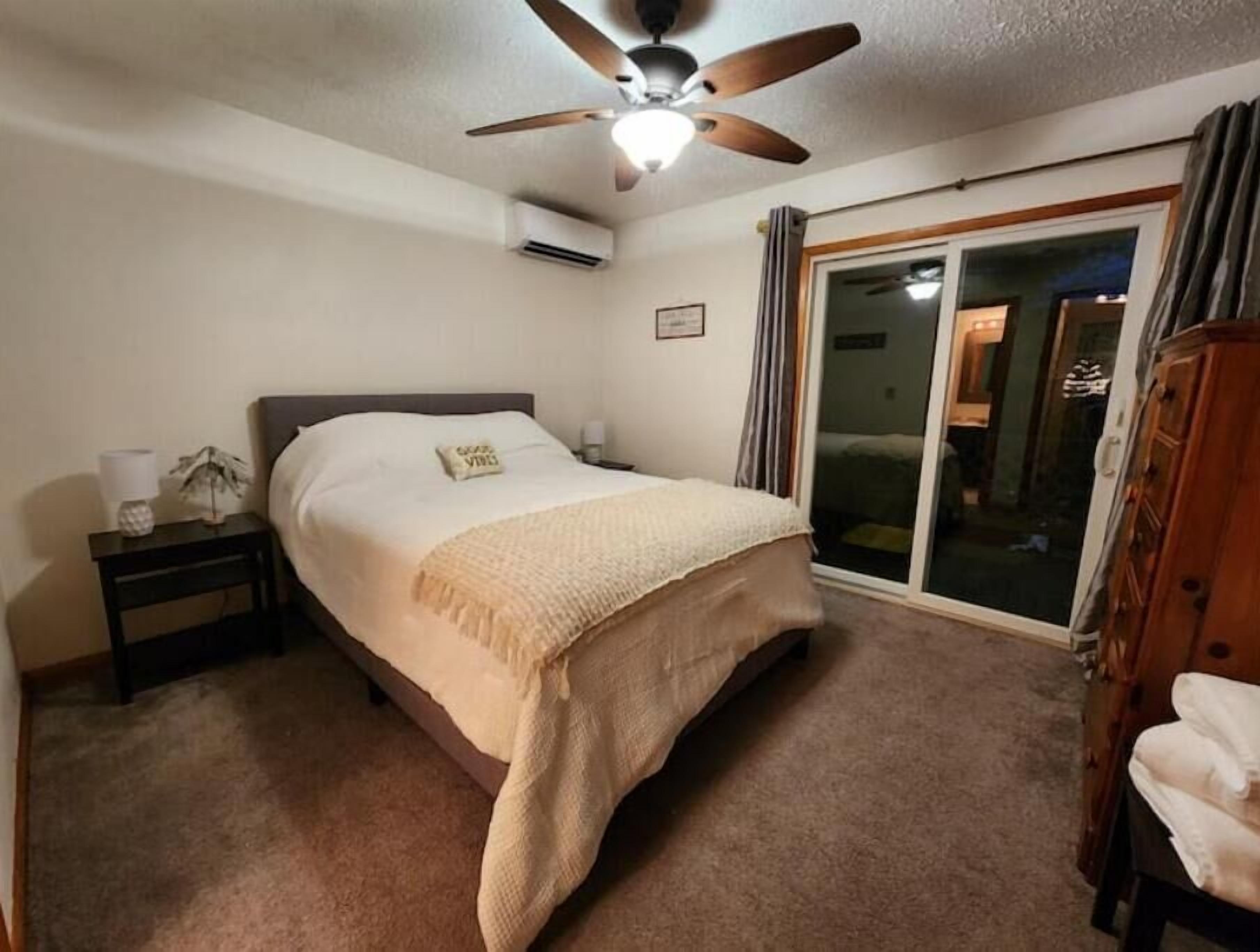 Ein Schlafzimmer mit einem Bett, einem Nachttisch, einem Deckenventilator, einer Klimaanlage, einer Glastür zum Verschieben und einem Holzschrank.