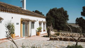 A white building with a tiled roof, a chimney, and a small balcony. There are potted plants and a stone pathway leading to the entrance. - Alojamiento Rural Para Grupos DE 20 Personas (BARBATE)