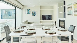 Un comedor moderno con una mesa blanca, sillas transparentes y un televisor al fondo.