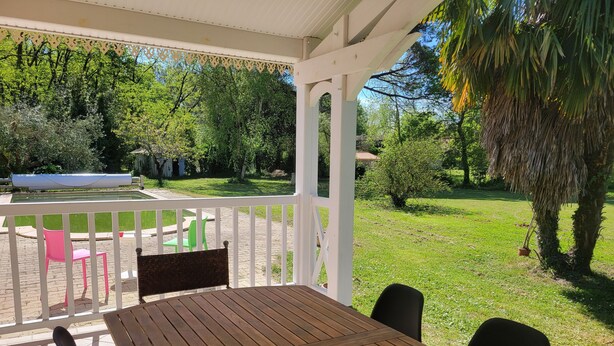 Een houten tafel met stoelen onder een witte pergola, met uitzicht op een gazon en bomen.