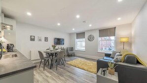 A modern living room with a sofa, a dining table with chairs, a TV, and a clock on the wall. - Central Windsor 1BR | Steps from Attractions (Windsor)