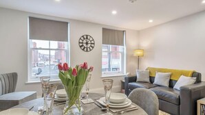 A modern living room with a dining area, large windows, a clock, and a sofa with cushions. - Central Windsor 1BR | Steps from Attractions (Windsor)