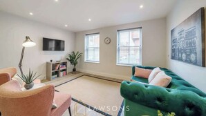 Una sala de estar con un sofá verde, un sillón rosa, una lámpara de pie, una estantería y un televisor montado en la pared.