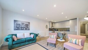 Una sala de estar moderna con un sofá color turquesa, un sillón rosa y una mesa de centro. Se ve una cocina al fondo con armarios blancos y un comedor.