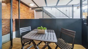 Un pequeño patio con una mesa, dos sillas, una planta en maceta y una escalera que sube a un nivel superior.