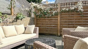 Een terras met een zithoek, inclusief een bank en rieten stoelen, een houten hek en een bakstenen muur.