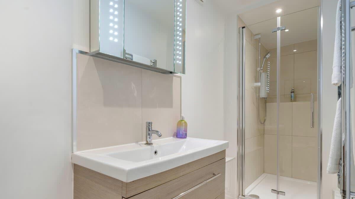 A modern bathroom with a white sink, a mirror with lights, a glass shower enclosure, and a wooden vanity.