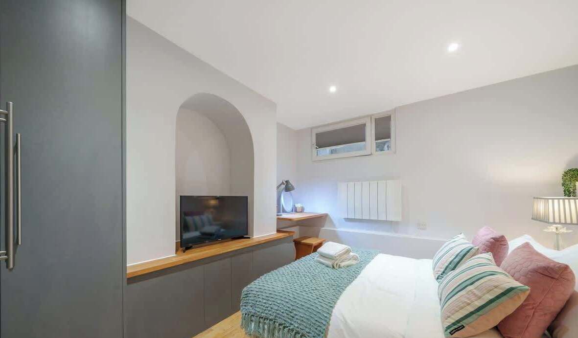 A modern bedroom with a TV on a wooden console, a bedside table with a lamp, a bed with pillows, and a window with a view.
